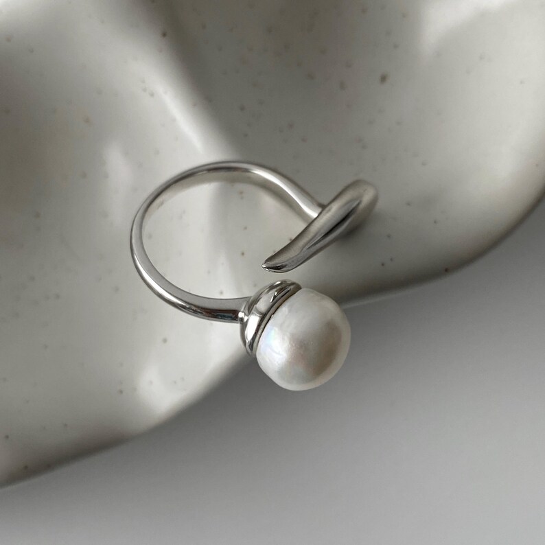 derMomentø - Handcrafted large pearl open ring with a naturally grown freshwater baroque pearl, set on a white rhodium-plated silver band. Display on a ceramic tray on a clean background.