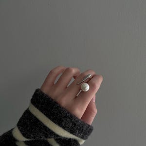 May include: A silver ring with a large white pearl on the top. The ring is worn on the middle finger of a hand.