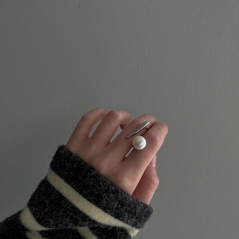 May include: A silver ring with a large white pearl on the top. The ring is worn on the middle finger of a hand.