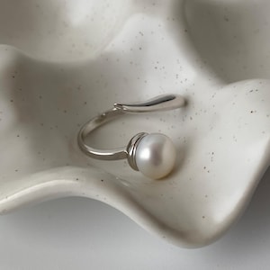 May include: A silver ring with a pearl and a curved band. The ring is on a white surface.