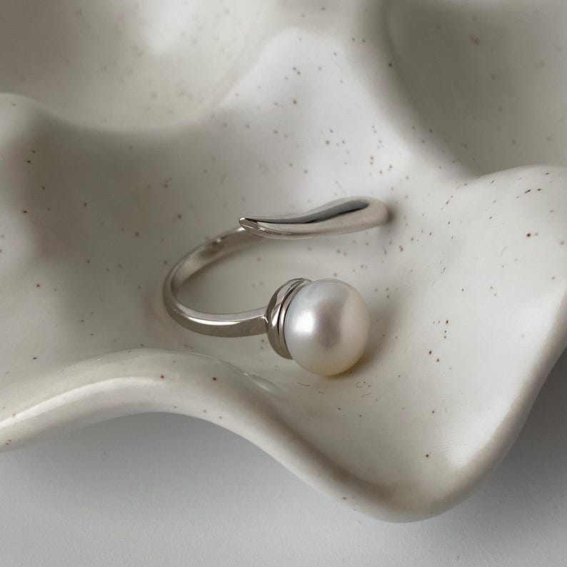 May include: A silver ring with a pearl and a curved band. The ring is on a white surface.
