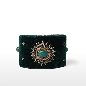 May include: A dark green velvet cuff bracelet with a central oval-shaped green stone surrounded by a silver-colored sunburst design. The bracelet has small silver-colored star-like accents and green beads on each side.