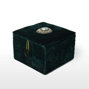 May include: A dark green velvet jewelry box with a silver and gemstone embellishment on the lid. The box has a gold-colored clasp and a small, round, black closure. The box is square-shaped and has a soft, textured appearance.