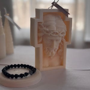 Könnte beinhalten: Eine weiße Kerze in Form eines Kreuzes mit einem detaillierten Bild von Jesu Christi Gesicht. Die Kerze hat ein handgemachtes Etikett und wird mit einem schwarzen Perlenarmband mit einem silbernen Kreuzanhänger präsentiert.