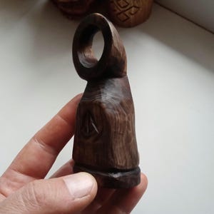 Hand holding a small, dark wooden figurine with a circular top and a carved triangular symbol on its body, likely a Slavic Panka doll or amulet.