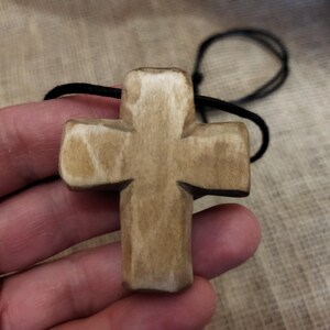 May include: A carved wooden cross pendant with a natural, light brown finish. The cross is suspended from a black cord. The pendant is held in a hand, showcasing the texture and detail of the wood.