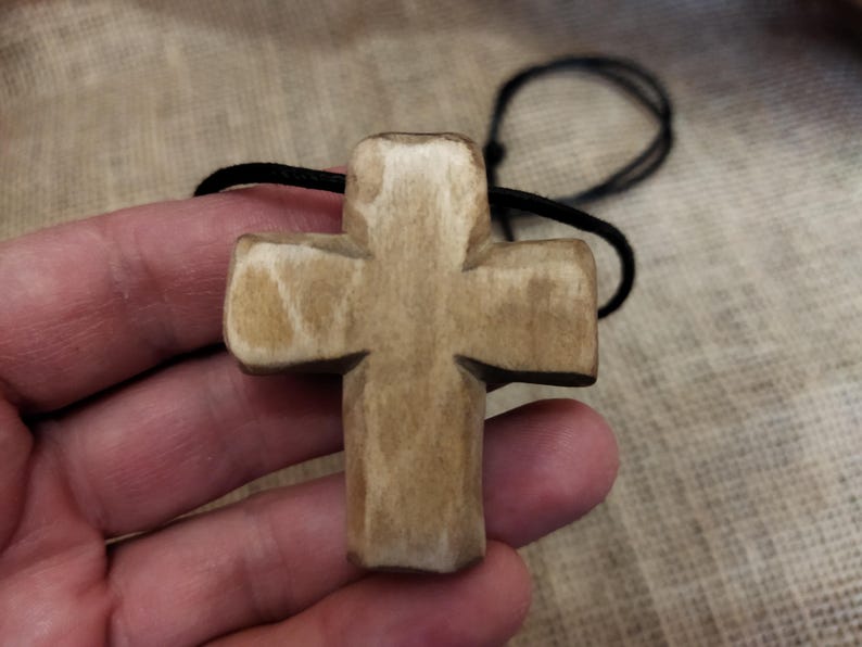 May include: A carved wooden cross pendant with a natural, light brown finish. The cross is suspended from a black cord. The pendant is held in a hand, showcasing the texture and detail of the wood.