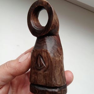 Hand holding a small, dark wooden figurine with a circular top and a carved triangular symbol on its body, likely a Slavic Panka doll or amulet.