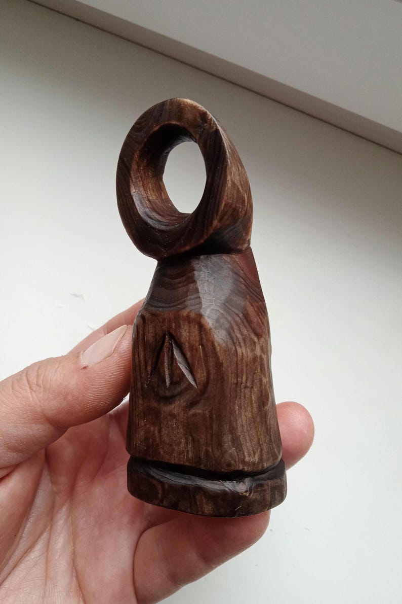 Hand holding a small, dark wooden figurine with a circular top and a carved triangular symbol on its body, likely a Slavic Panka doll or amulet.