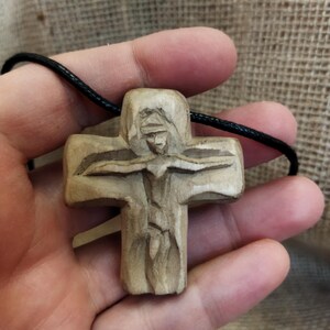 May include: A carved wooden pendant depicting a figure on a cross. The pendant is beige and hangs from a black cord. The carving shows the figure's arms outstretched. The background is a textured, neutral color.