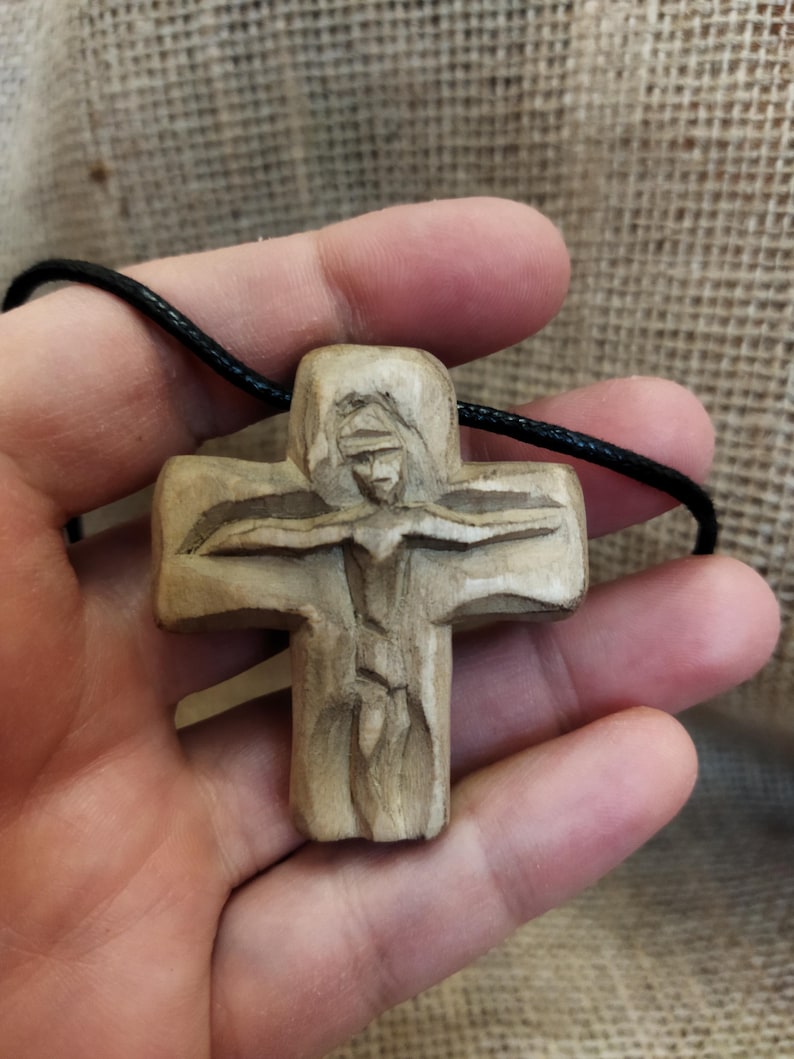 May include: A carved wooden pendant depicting a figure on a cross. The pendant is beige and hangs from a black cord. The carving shows the figure's arms outstretched. The background is a textured, neutral color.