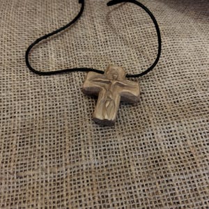 May include: A wooden cross pendant with a carved figure of Jesus on a black cord. The cross is light brown and rests on a textured, tan fabric background. The necklace is a religious item.