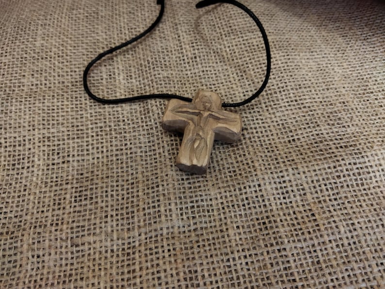 May include: A wooden cross pendant with a carved figure of Jesus on a black cord. The cross is light brown and rests on a textured, tan fabric background. The necklace is a religious item.
