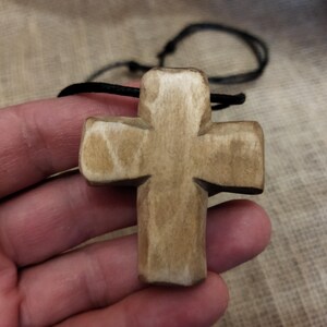 May include: A wooden cross pendant with a natural finish, suspended on a black cord. The cross is held in a hand, showcasing its carved details and the texture of the wood. The background is a neutral fabric.