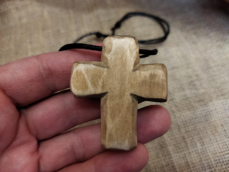 May include: A wooden cross pendant with a natural finish, suspended on a black cord. The cross is held in a hand, showcasing its carved details and the texture of the wood. The background is a neutral fabric.