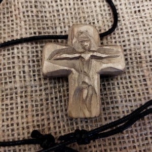May include: A carved wooden cross pendant with a detailed depiction of a figure. The cross is attached to a black cord. The background is a textured, woven fabric.