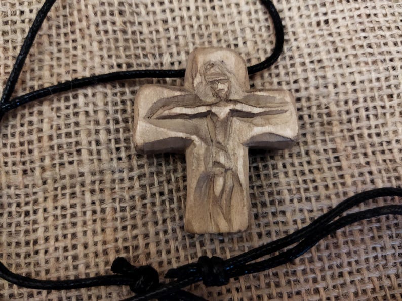 May include: A carved wooden cross pendant with a detailed depiction of a figure. The cross is attached to a black cord. The background is a textured, woven fabric.