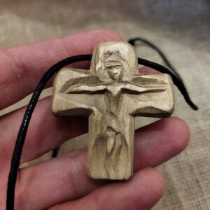 May include: A carved wooden cross pendant with a detailed figure. The cross is light brown and hangs from a black cord. The carving depicts a figure with outstretched arms. The background is a neutral tone.