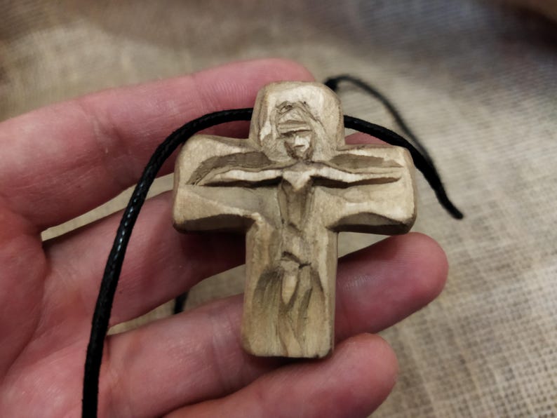 May include: A carved wooden cross pendant with a detailed figure. The cross is light brown and hangs from a black cord. The carving depicts a figure with outstretched arms. The background is a neutral tone.