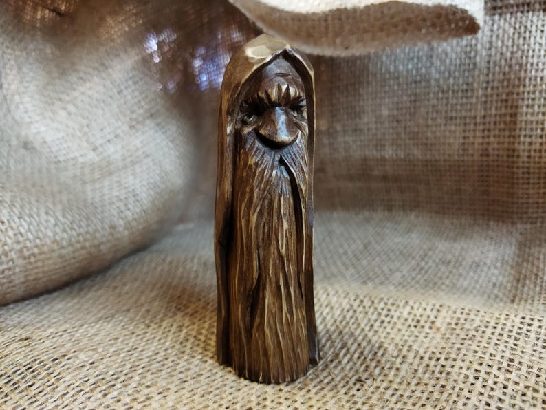 A 3.6-inch tall hand-carved linden wood figurine of a forest spirit. The figure has a long, flowing beard with deep textures and a face partially hidden under a hood. The dark brown stain highlights the artisan's hand-cut marks. This unique piece of Ukrainian folk art has a matte beeswax finish and a rustic, mystical appearance, shown against a neutral background.