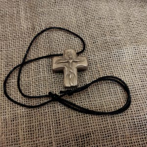 May include: A carved wooden cross pendant with a black cord necklace. The cross features a detailed depiction of a figure. The necklace is lying on a textured, tan fabric.