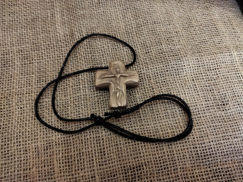 May include: A carved wooden cross pendant with a black cord necklace. The cross features a detailed depiction of a figure. The necklace is lying on a textured, tan fabric.