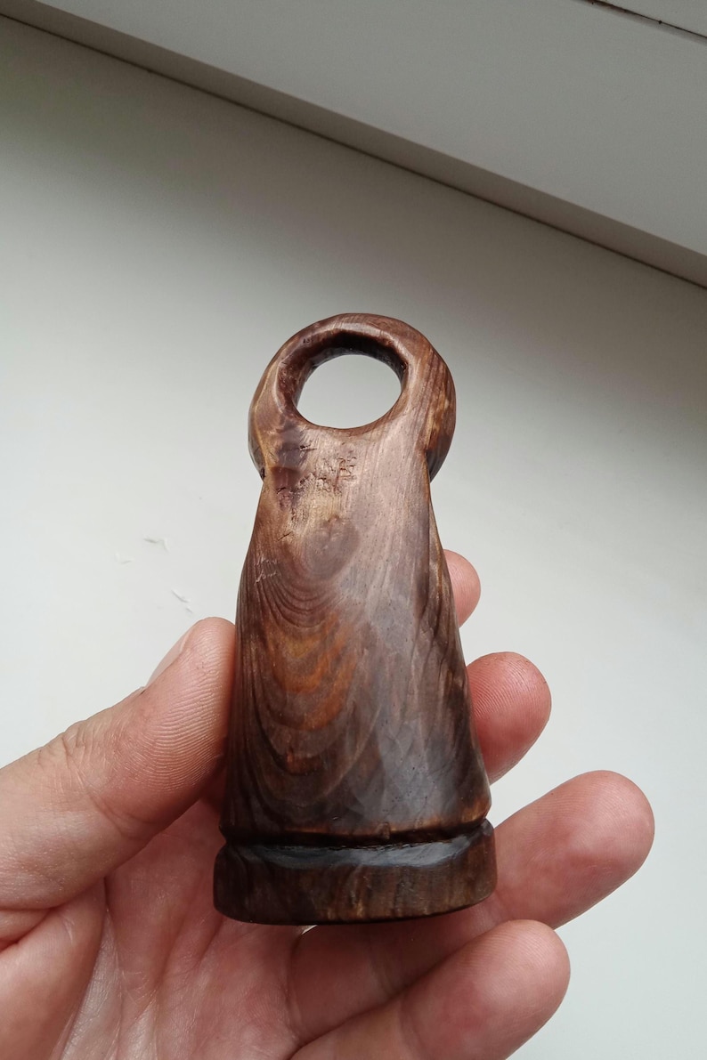 Hand holding a small, dark wooden figurine with a circular top and a carved triangular symbol on its body, likely a Slavic Panka doll or amulet.