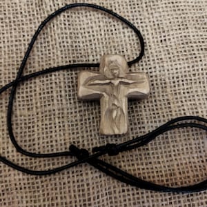 May include: A wooden cross pendant with a carved figure, suspended on a black cord. The cross is light brown, and the cord is knotted. The background is a textured, tan fabric.
