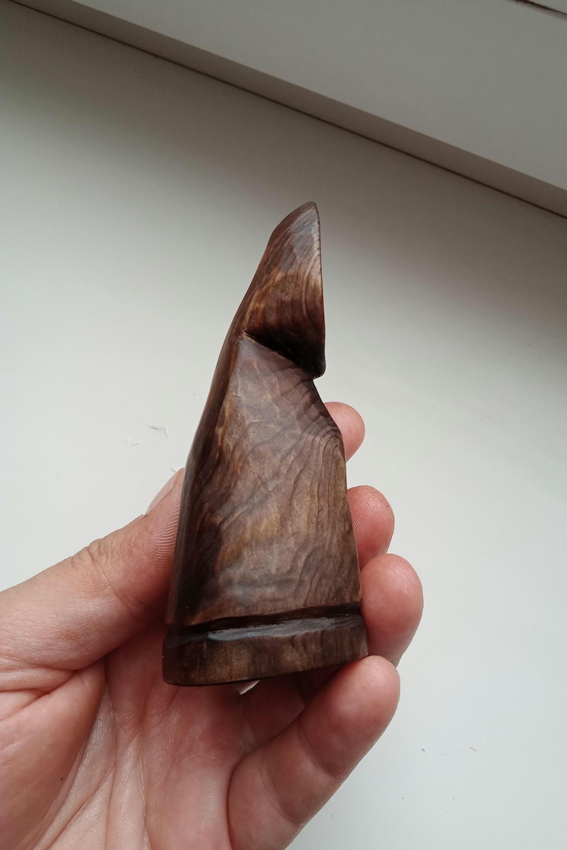 Hand holding a small, dark wooden figurine with a circular top and a carved triangular symbol on its body, likely a Slavic Panka doll or amulet.