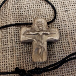 May include: A carved wooden cross pendant with a detailed depiction of a figure. The cross is attached to a black cord. The pendant is made of light-colored wood and has a natural finish. The background is a textured, tan fabric.