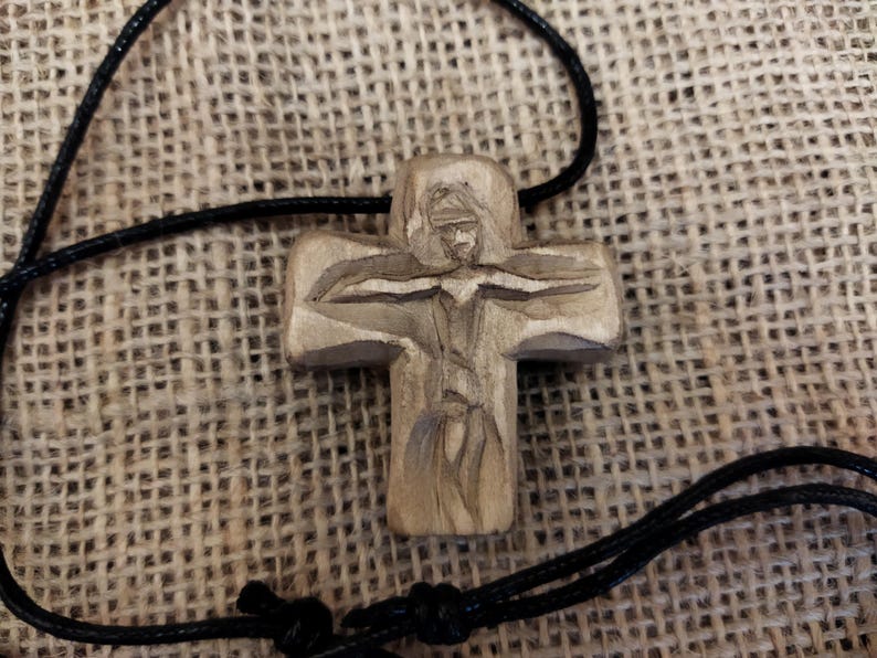 May include: A carved wooden cross pendant with a detailed depiction of a figure. The cross is attached to a black cord. The pendant is made of light-colored wood and has a natural finish. The background is a textured, tan fabric.