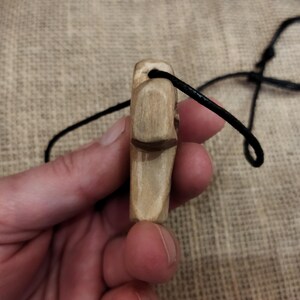 May include: A hand-carved wooden pendant with a dark brown accent and a black cord. The pendant is light brown and rectangular, with a carved design. The cord is knotted at the top.