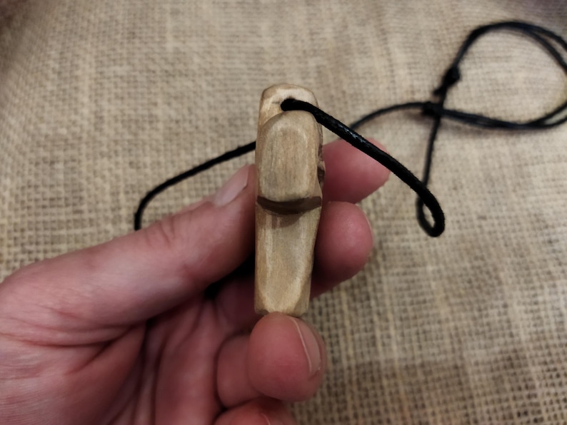 May include: A hand-carved wooden pendant with a dark brown accent and a black cord. The pendant is light brown and rectangular, with a carved design. The cord is knotted at the top.