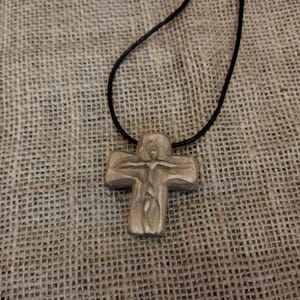 May include: A wooden cross pendant on a black cord necklace. The cross is carved with a figure and has a natural wood grain finish. The necklace rests on a textured, tan fabric background.