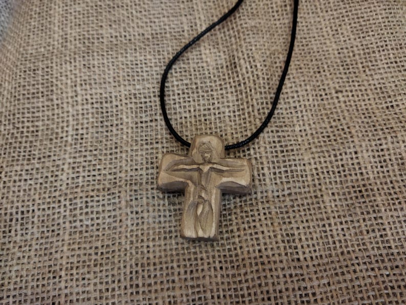 May include: A wooden cross pendant on a black cord necklace. The cross is carved with a figure and has a natural wood grain finish. The necklace rests on a textured, tan fabric background.