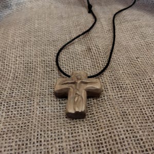 May include: A wooden cross pendant on a black cord. The cross is carved with a figure and has a natural wood color. The necklace is displayed against a textured, neutral-toned background.