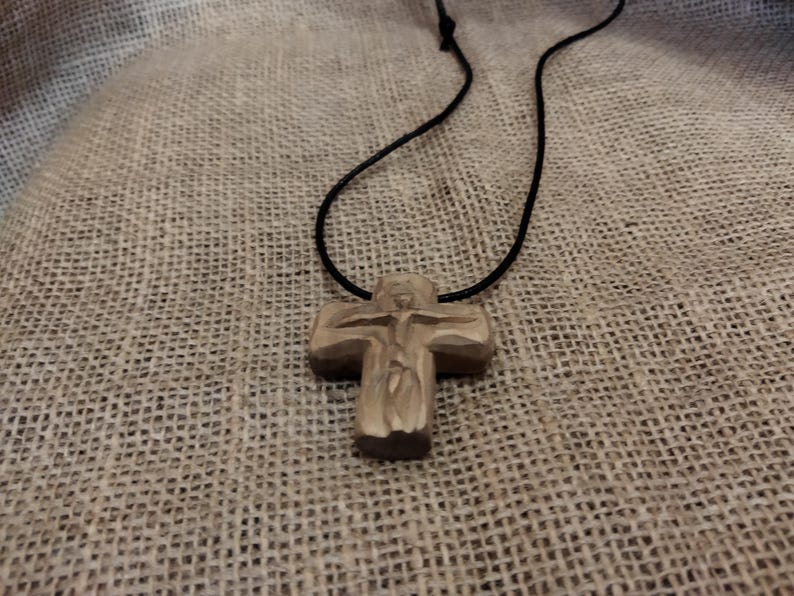 May include: A wooden cross pendant on a black cord. The cross is carved with a figure and has a natural wood color. The necklace is displayed against a textured, neutral-toned background.