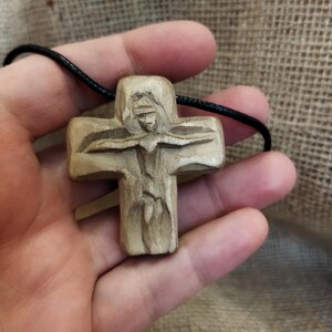 May include: A carved wooden cross pendant on a black cord. The cross features a detailed depiction of a figure, likely religious, with outstretched arms. The wood has a natural, light brown color, and the carving shows intricate details.