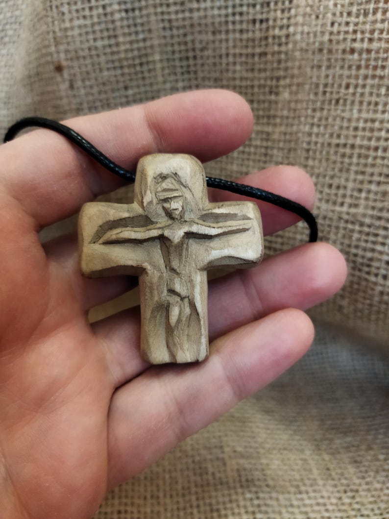 May include: A carved wooden cross pendant on a black cord. The cross features a detailed depiction of a figure, likely religious, with outstretched arms. The wood has a natural, light brown color, and the carving shows intricate details.