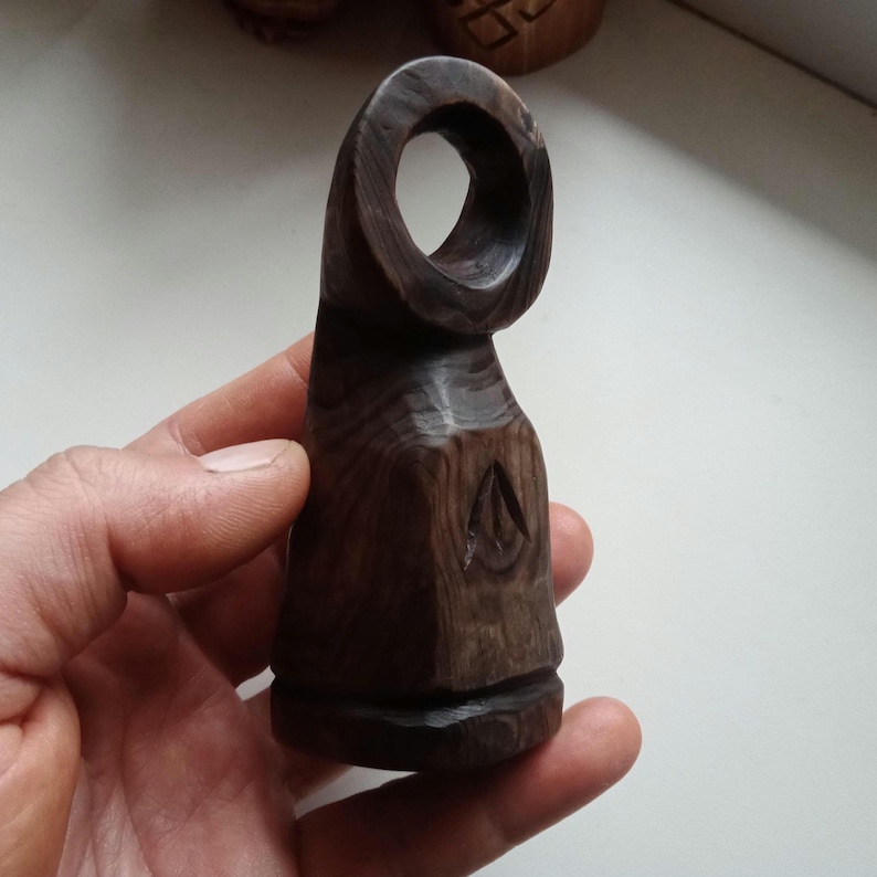 Hand holding a small, dark wooden figurine with a circular top and a carved triangular symbol on its body, likely a Slavic Panka doll or amulet.