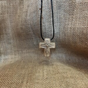 May include: A beige-colored cross pendant with a carved figure of Jesus Christ on a black cord. The cross is made of a light-colored material and is suspended against a textured, tan background.