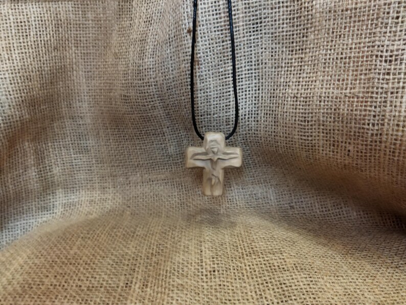 May include: A beige-colored cross pendant with a carved figure of Jesus Christ on a black cord. The cross is made of a light-colored material and is suspended against a textured, tan background.