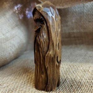 A 3.6-inch tall hand-carved linden wood figurine of a forest spirit. The figure has a long, flowing beard with deep textures and a face partially hidden under a hood. The dark brown stain highlights the artisan's hand-cut marks. This unique piece of Ukrainian folk art has a matte beeswax finish and a rustic, mystical appearance, shown against a neutral background.