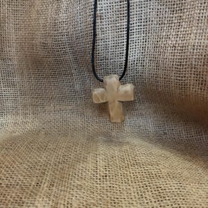 May include: A light-colored, cross-shaped pendant hangs from a black cord. The cross has a textured surface and is suspended against a textured, tan fabric background. The pendant is a religious symbol.
