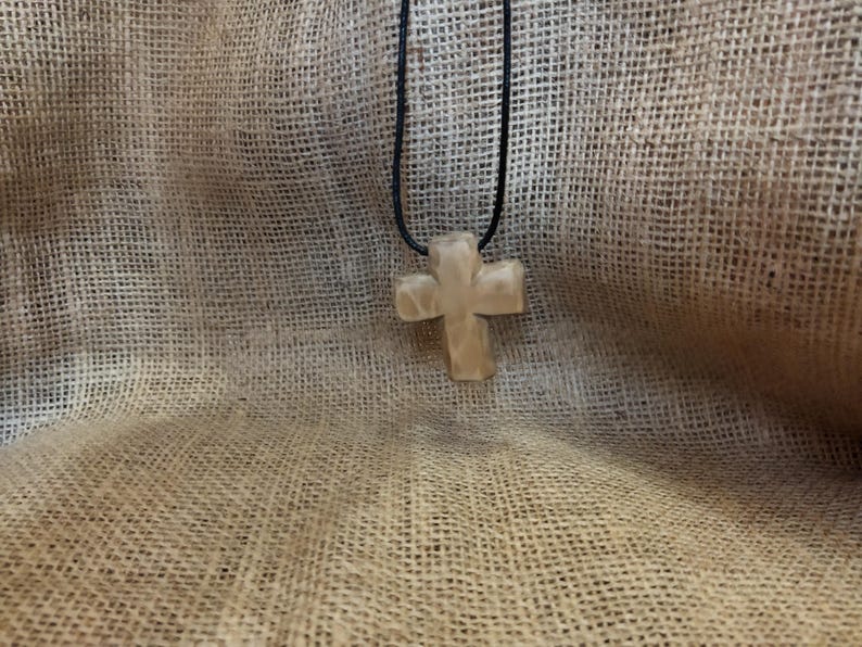May include: A light-colored, cross-shaped pendant hangs from a black cord. The cross has a textured surface and is suspended against a textured, tan fabric background. The pendant is a religious symbol.