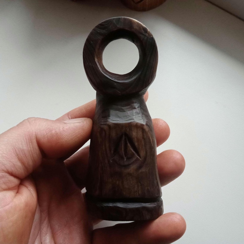 Hand holding a small, dark wooden figurine with a circular top and a carved triangular symbol on its body, likely a Slavic Panka doll or amulet.