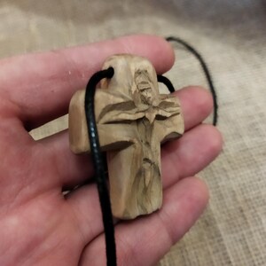 May include: A carved wooden cross pendant with a detailed figure, suspended on a black cord. The cross is light brown, with visible wood grain and intricate carvings. The cord is thin and black, suitable for wearing as a necklace.