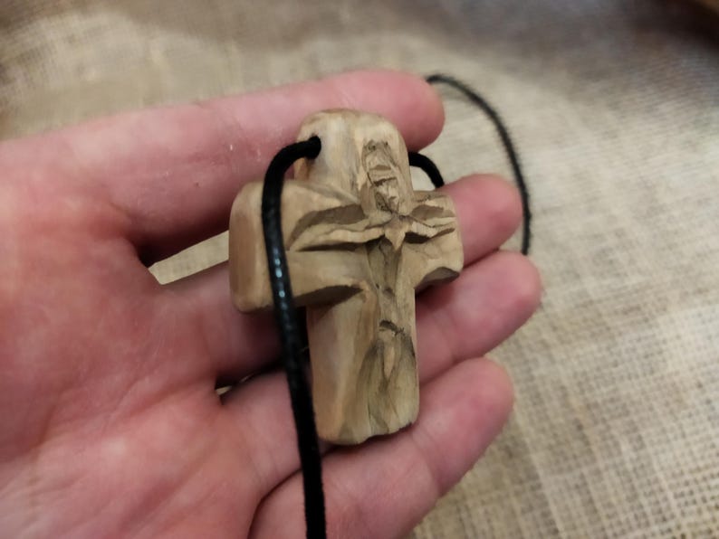May include: A carved wooden cross pendant with a detailed figure, suspended on a black cord. The cross is light brown, with visible wood grain and intricate carvings. The cord is thin and black, suitable for wearing as a necklace.