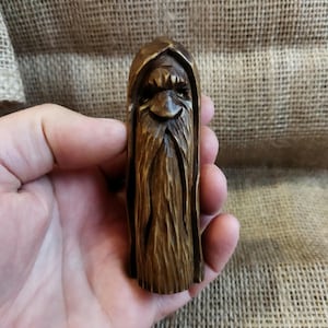 A 3.6-inch tall hand-carved linden wood figurine of a forest spirit. The figure has a long, flowing beard with deep textures and a face partially hidden under a hood. The dark brown stain highlights the artisan's hand-cut marks. This unique piece of Ukrainian folk art has a matte beeswax finish and a rustic, mystical appearance, shown against a neutral background.