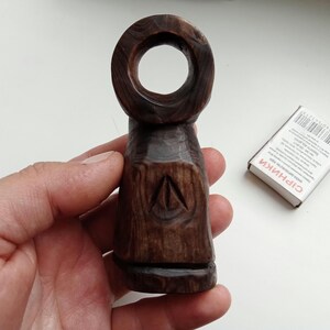 Hand holding a small, dark wooden figurine with a circular top and a carved triangular symbol on its body, likely a Slavic Panka doll or amulet.